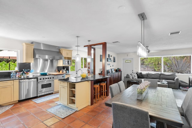 kitchen featuring a center island with sink, a kitchen breakfast bar, pendant lighting, stainless steel appliances, and wall chimney range hood