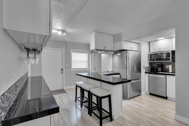 kitchen with appliances with stainless steel finishes, under cabinet range hood, a kitchen bar, tasteful backsplash, and light wood-style floors