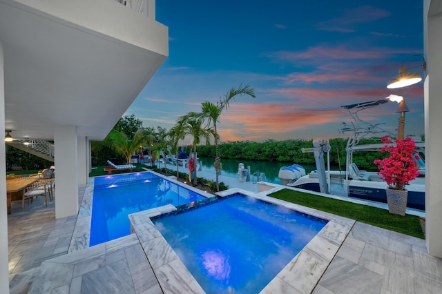 pool at dusk featuring an in ground hot tub, a water view, and a patio area