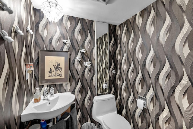 half bathroom featuring a sink, a notable chandelier, toilet, and wallpapered walls