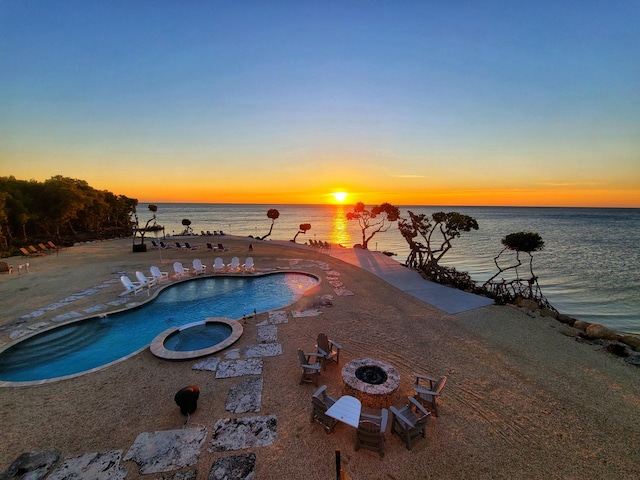 pool at dusk with a fire pit, an in ground hot tub, a patio, and a water view