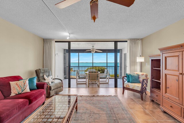 tiled living room featuring expansive windows, a water view, a textured ceiling, and ceiling fan