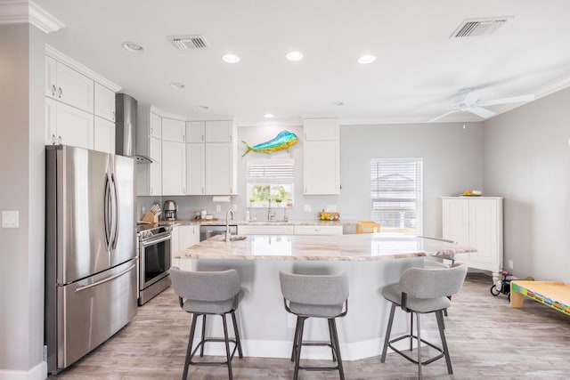 kitchen with white cabinets, a kitchen island with sink, light stone counters, stainless steel appliances, and wall chimney exhaust hood