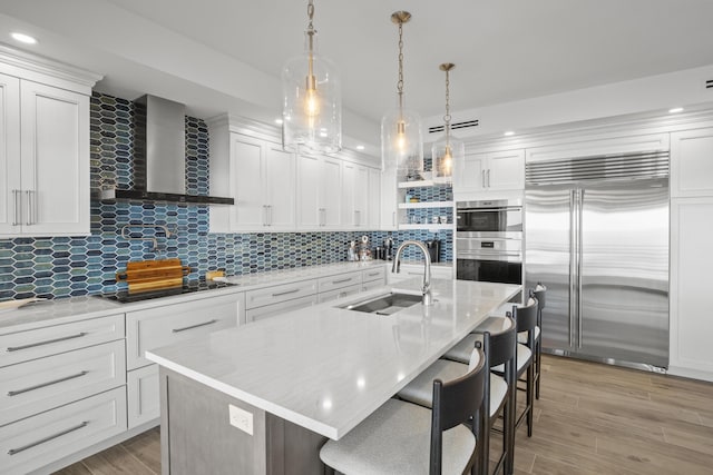 kitchen featuring appliances with stainless steel finishes, white cabinetry, sink, a center island with sink, and wall chimney exhaust hood