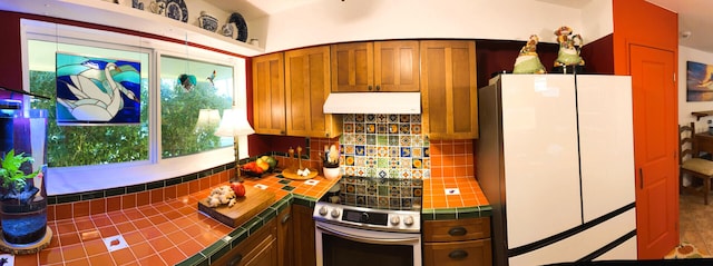 kitchen with tile countertops, freestanding refrigerator, under cabinet range hood, stainless steel electric range, and plenty of natural light