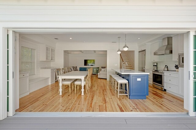 kitchen featuring wall chimney exhaust hood, built in appliances, ornamental molding, a sink, and light wood-style floors
