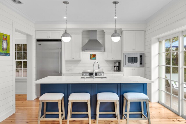 kitchen featuring wall chimney exhaust hood, built in appliances, white cabinets, an island with sink, and ornamental molding