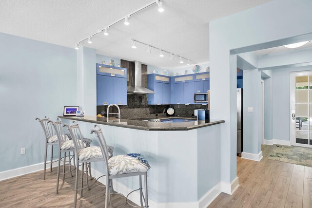 kitchen with a breakfast bar area, blue cabinetry, appliances with stainless steel finishes, kitchen peninsula, and wall chimney exhaust hood