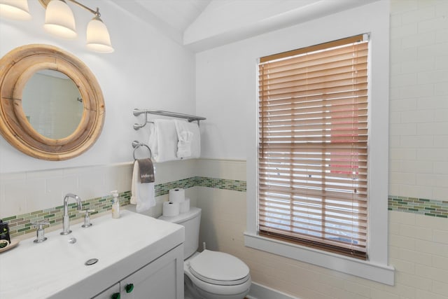 bathroom featuring a wainscoted wall, tile walls, toilet, vaulted ceiling, and vanity