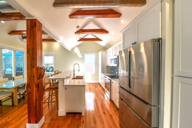 kitchen with a kitchen island with sink, light wood-style floors, lofted ceiling with beams, a sink, and appliances with stainless steel finishes