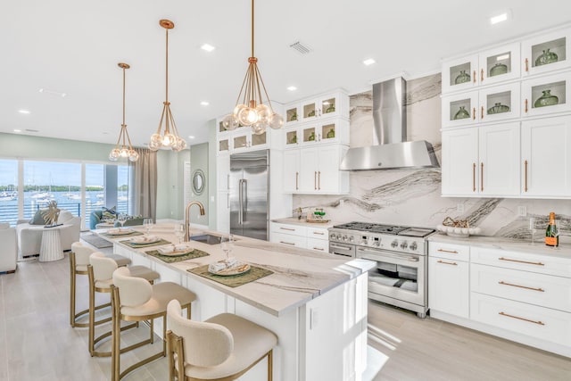 kitchen with pendant lighting, wall chimney range hood, high end appliances, light stone countertops, and a center island with sink