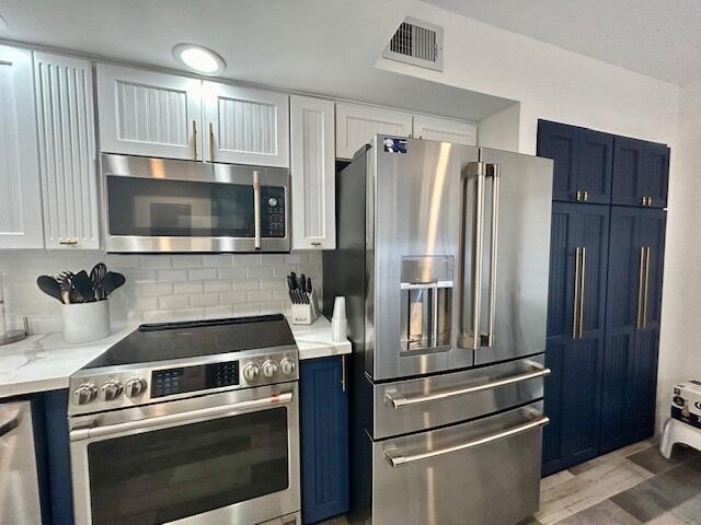 kitchen with appliances with stainless steel finishes, blue cabinets, white cabinetry, tasteful backsplash, and light stone countertops