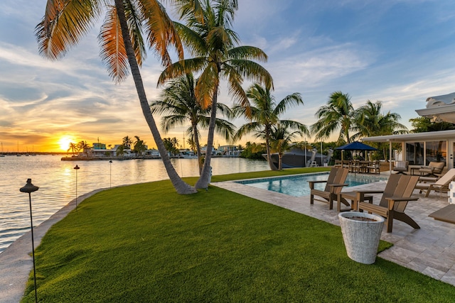 pool at dusk with a patio area, a lawn, and a water view