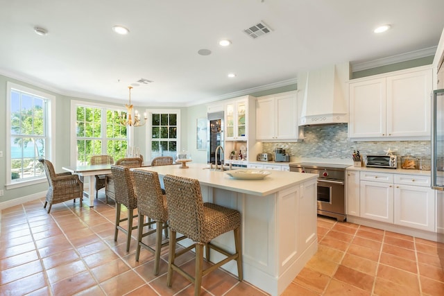 kitchen featuring high end stainless steel range, custom exhaust hood, hanging light fixtures, a kitchen island with sink, and backsplash