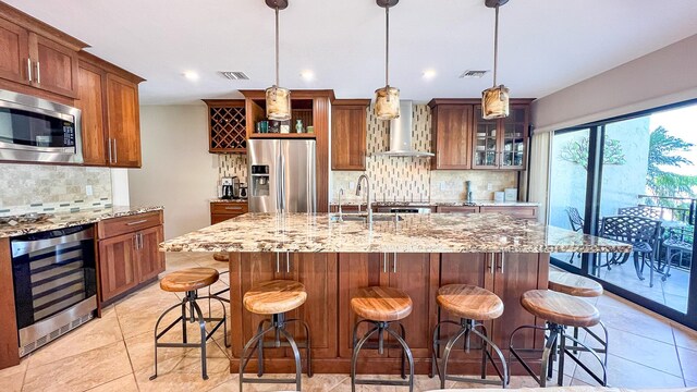 kitchen featuring appliances with stainless steel finishes, beverage cooler, hanging light fixtures, a kitchen island with sink, and wall chimney range hood