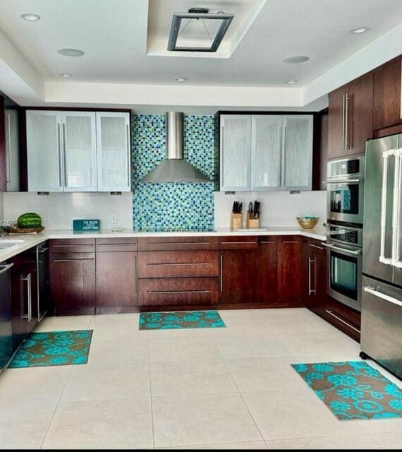 kitchen with light tile patterned flooring, stainless steel appliances, light countertops, wall chimney range hood, and backsplash