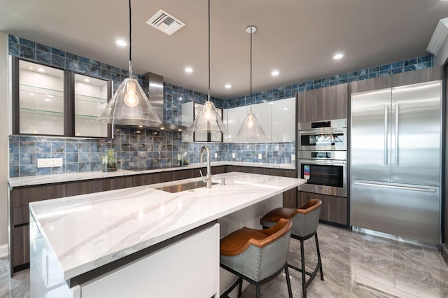 kitchen featuring appliances with stainless steel finishes, an island with sink, decorative backsplash, hanging light fixtures, and wall chimney range hood