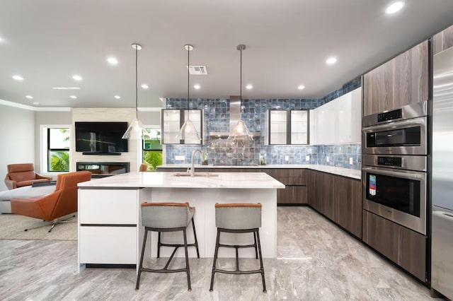 kitchen featuring sink, wall chimney range hood, a kitchen island with sink, backsplash, and a kitchen breakfast bar