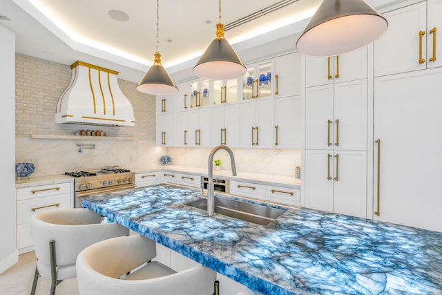 kitchen featuring high end stainless steel range, sink, custom exhaust hood, a tray ceiling, and white cabinets