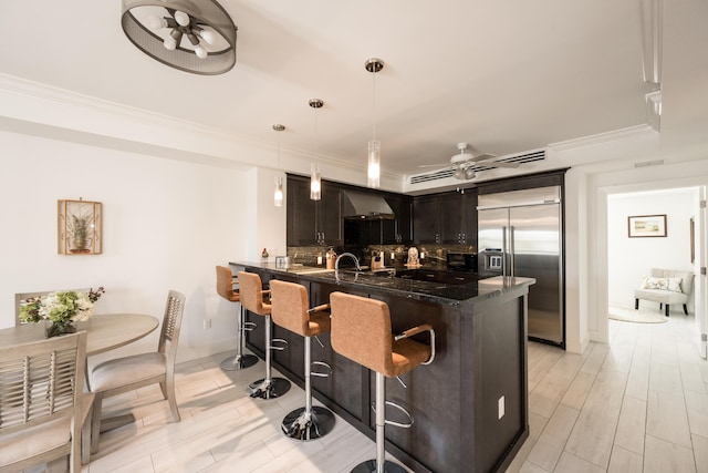 kitchen featuring a breakfast bar, pendant lighting, built in refrigerator, kitchen peninsula, and wall chimney exhaust hood