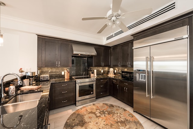 kitchen with sink, dark brown cabinets, appliances with stainless steel finishes, dark stone counters, and wall chimney range hood