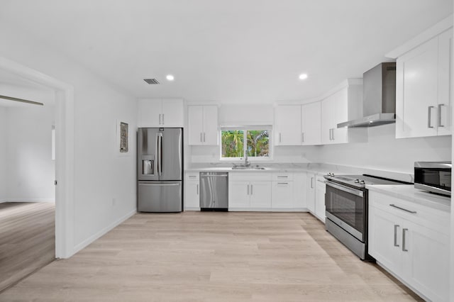 kitchen featuring appliances with stainless steel finishes, wall chimney exhaust hood, a sink, white cabinets, and light wood-type flooring