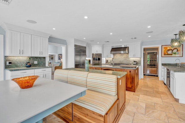 kitchen featuring wall chimney range hood, appliances with stainless steel finishes, hanging light fixtures, white cabinets, and a center island with sink