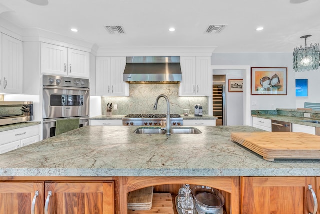 kitchen with decorative light fixtures, white cabinetry, stainless steel appliances, a center island with sink, and wall chimney exhaust hood