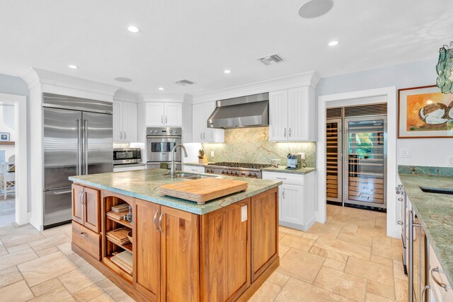 kitchen featuring white cabinetry, wall chimney exhaust hood, stainless steel appliances, and a kitchen island with sink