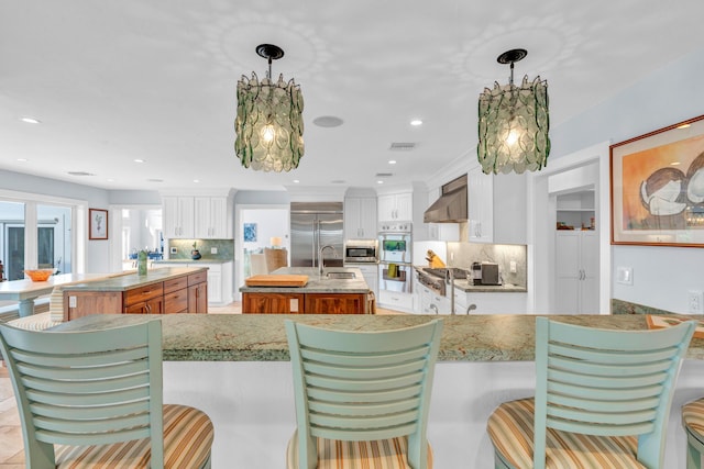 kitchen featuring wall chimney range hood, a center island with sink, white cabinets, and decorative light fixtures