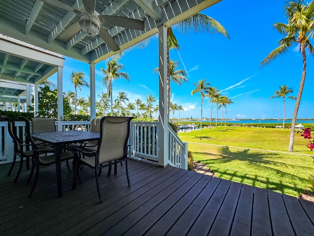wooden terrace with outdoor dining space, ceiling fan, a water view, and a yard