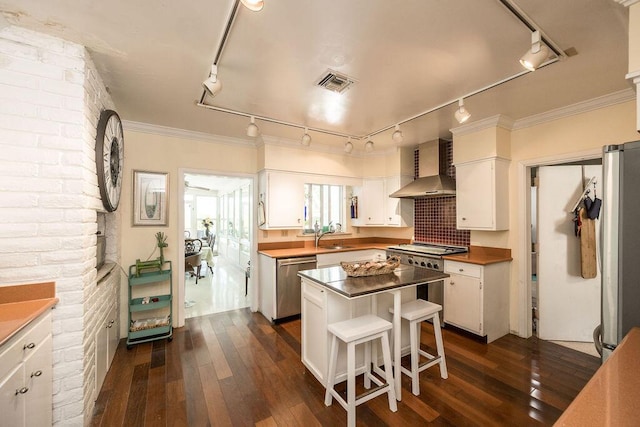 kitchen with wall chimney range hood, dark wood-type flooring, appliances with stainless steel finishes, white cabinetry, and a kitchen island