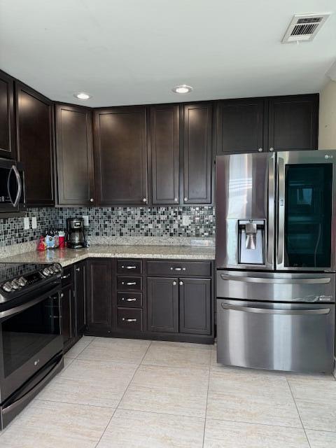 kitchen with appliances with stainless steel finishes, dark brown cabinets, light stone counters, light tile patterned flooring, and decorative backsplash