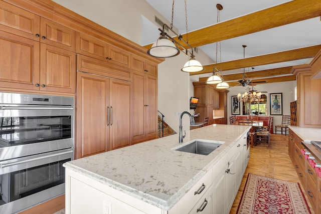 kitchen with stainless steel double oven, a kitchen island with sink, a sink, beam ceiling, and decorative light fixtures