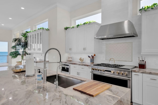 kitchen with wall chimney exhaust hood, light stone counters, plenty of natural light, double oven range, and white cabinets