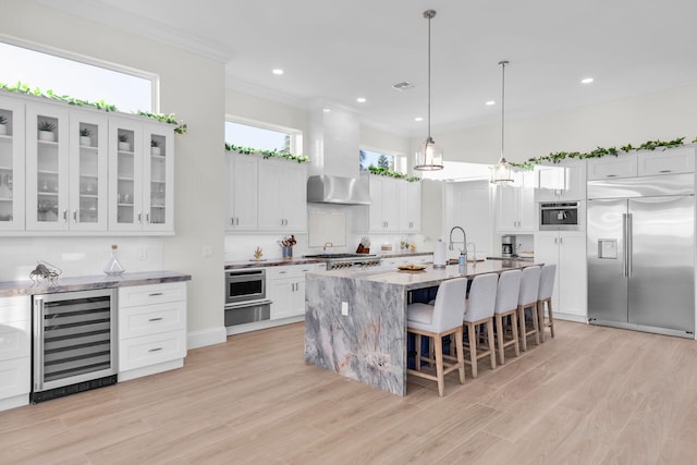 kitchen featuring white cabinetry, built in appliances, wine cooler, a center island with sink, and wall chimney exhaust hood