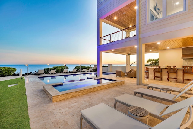 pool at dusk with an outdoor bar, a patio, a water view, an outdoor living space, and an in ground hot tub