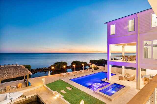 pool at dusk with exterior kitchen, a water view, a bar, an outdoor living space with a fire pit, and an in ground hot tub