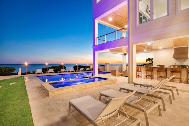 pool at dusk featuring an in ground hot tub, a water view, exterior bar, and a patio area