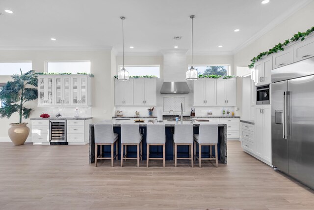 kitchen featuring pendant lighting, wall chimney range hood, white cabinetry, a kitchen island with sink, and stainless steel built in fridge