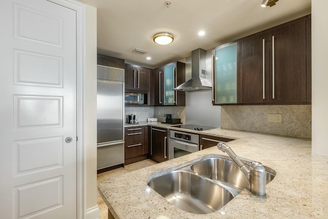 kitchen featuring appliances with stainless steel finishes, a sink, dark brown cabinets, light stone countertops, and wall chimney exhaust hood