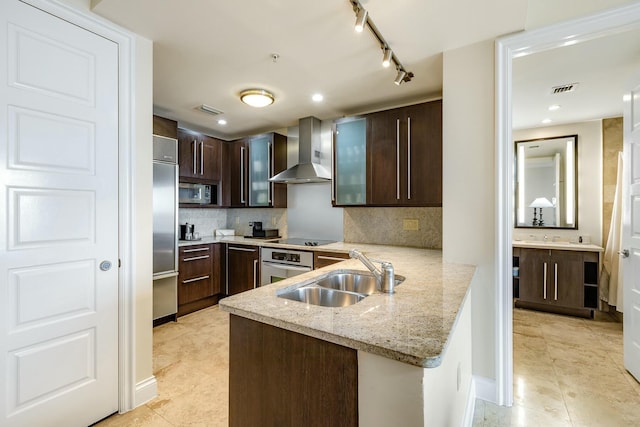 kitchen featuring a sink, light stone countertops, a peninsula, wall chimney exhaust hood, and built in appliances