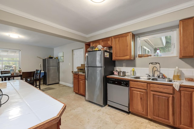 kitchen with a sink, appliances with stainless steel finishes, tile counters, brown cabinetry, and crown molding