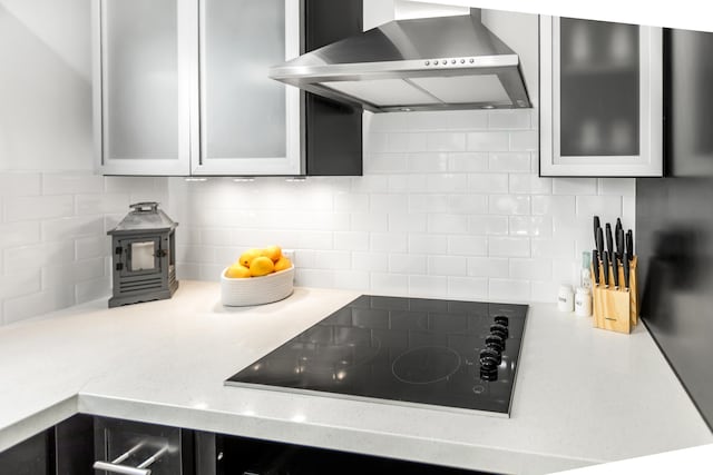 kitchen featuring black electric stovetop, light countertops, backsplash, glass insert cabinets, and wall chimney exhaust hood