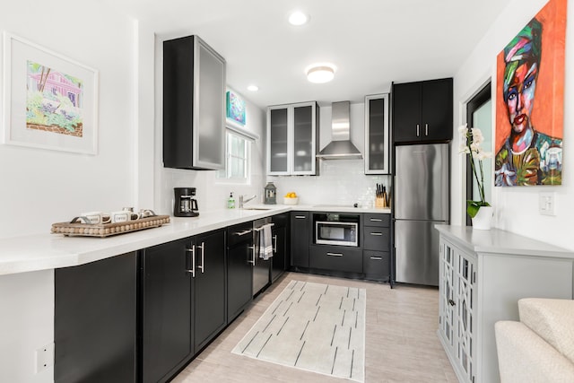 kitchen featuring light countertops, dark cabinetry, freestanding refrigerator, wall chimney exhaust hood, and glass insert cabinets