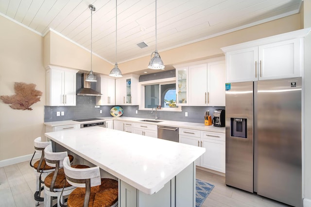 kitchen with wall chimney exhaust hood, hanging light fixtures, appliances with stainless steel finishes, a kitchen island, and decorative backsplash