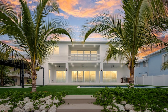 back house at dusk with a patio area, a balcony, a yard, ceiling fan, and a swimming pool