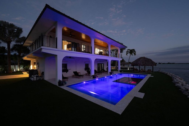 pool at dusk featuring an in ground hot tub, ceiling fan, a water view, and a patio area