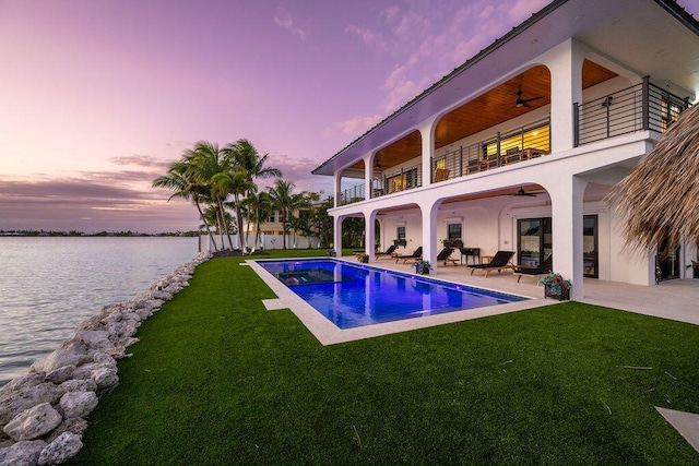 pool at dusk featuring a patio, a water view, ceiling fan, and a lawn