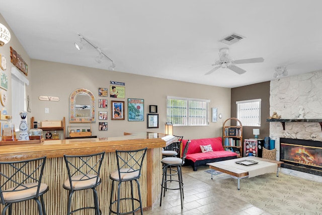 living room featuring rail lighting, a stone fireplace, bar, and ceiling fan